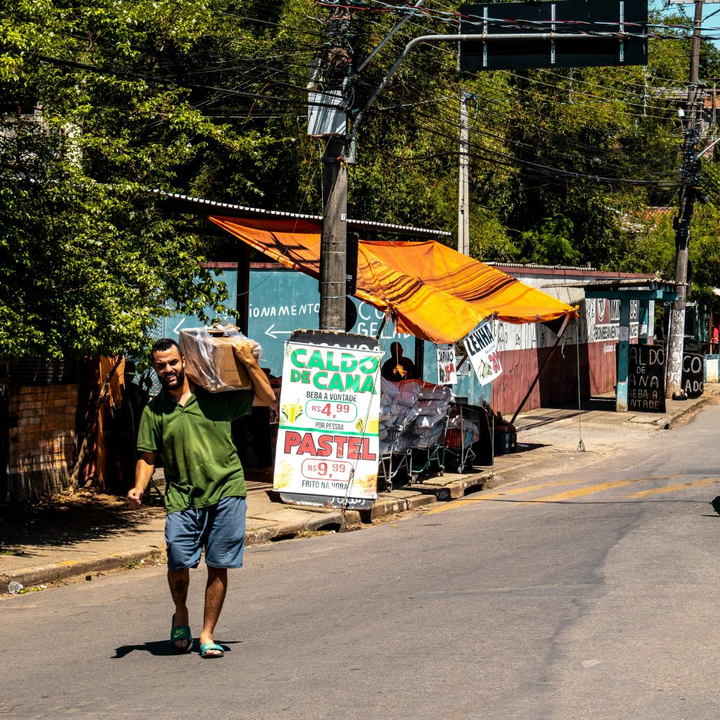 Smiling man carries package through vibrant street, past food vendors and tangled wires.